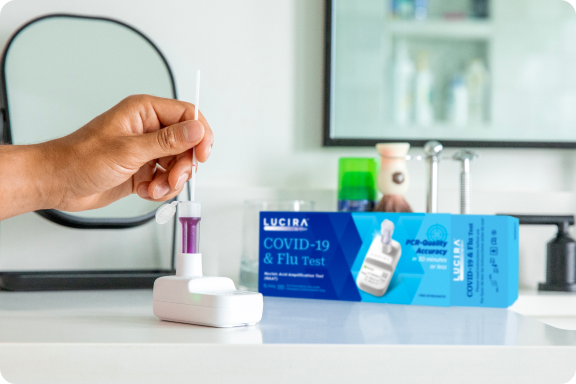 A hand comes in and uses the LUCIRA® by Pfizer COVID-19 & Flu Test with the packaging sitting on the right on top of the counter.