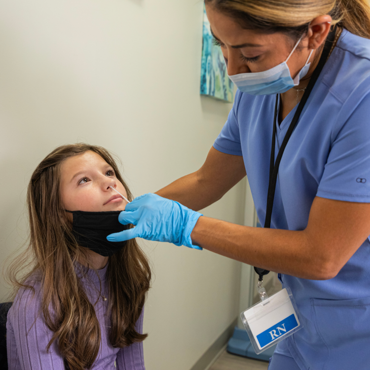 A healthcare professional is wearing PPE and using a swab on a young patient with a lowered mask to test for COVID-19 and the flu.