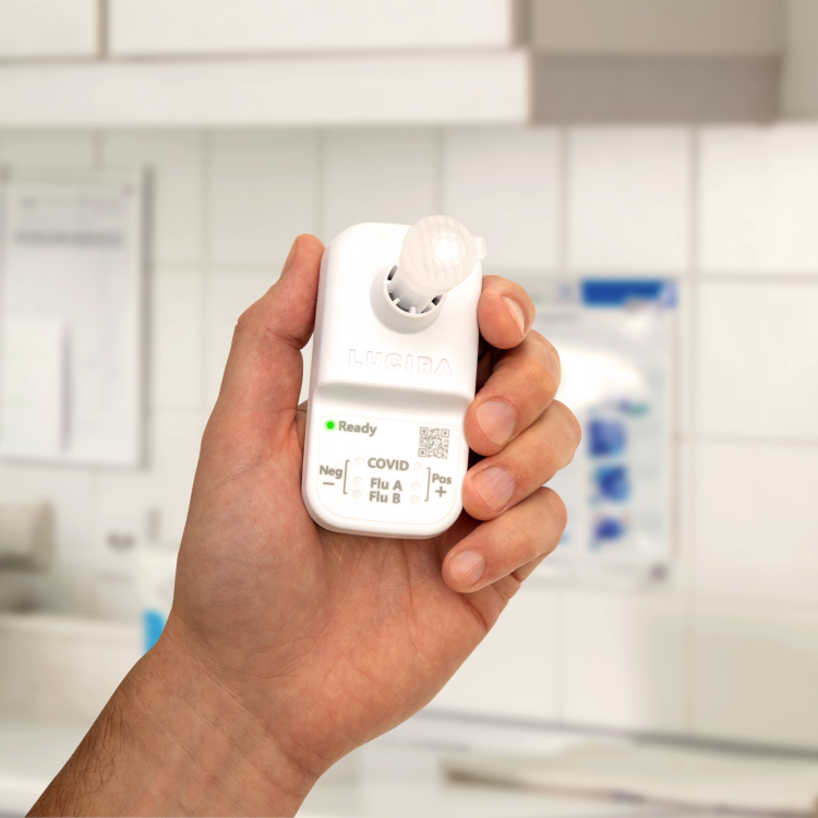 A hand holds up the the LUCIRA® by Pfizer COVID-19 & Flu Test, a small white device.