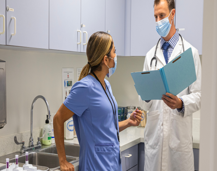 A doctor and physician talk to one another with masks on in a medical room.