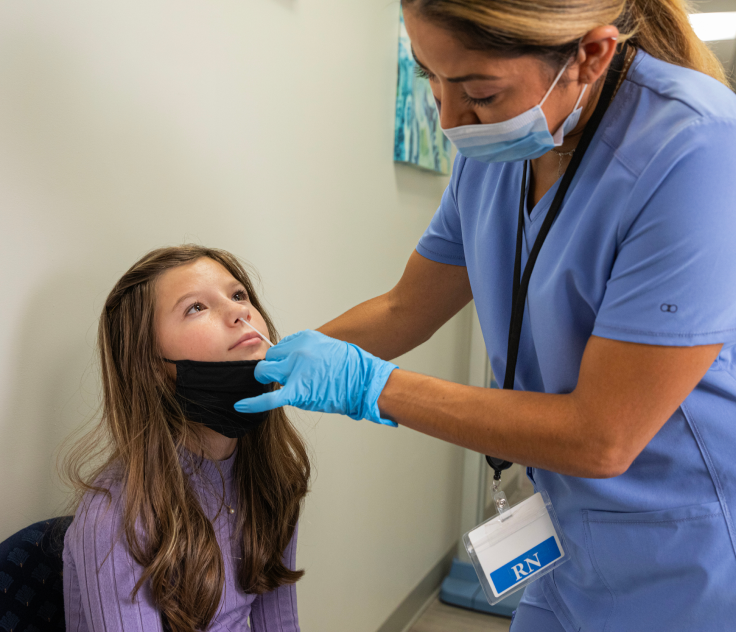 A healthcare professional is wearing PPE and using a swab on a young patient with a lowered mask to test for COVID-19 and the flu.