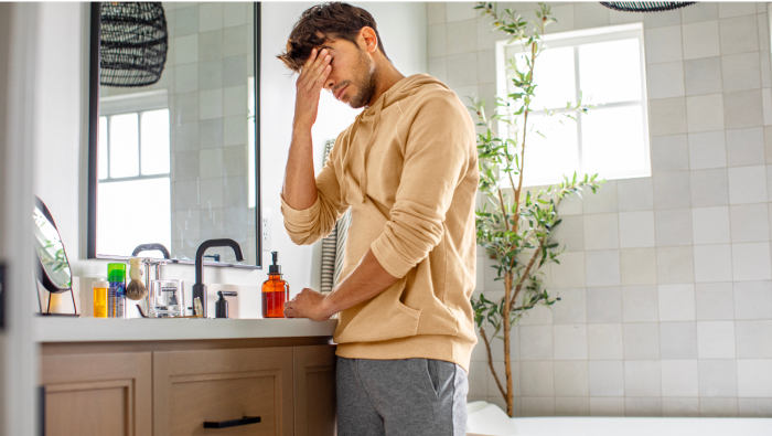 An image of a worried man in his bathroom. His hand is pressed to his head.