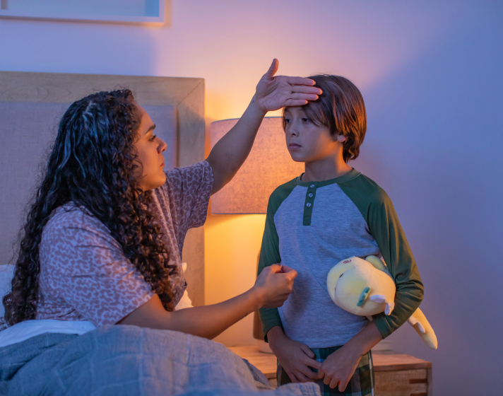 An image of a mom sitting on the bed as her child stands in front of her. Her hand is on the child's head to check his temperature.