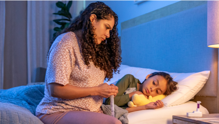 An image of a worried mom sitting next to her child in bed. The LUCIRA® by Pfizer COVID-19 & Flu Home Test sits on the bedside table.