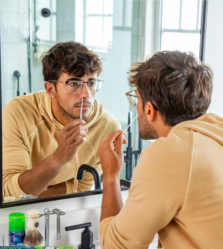 An image of a man with glasses as he administers a nasal swab in his bathroom while looking in the mirror.