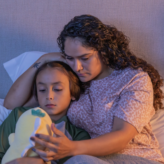 An image of a concerned mom lying in bed with her child, as they look at a stuffed animal.