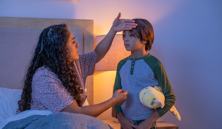 An image of a mom sitting on the bed as her child stands in front of her. Her hand is on the child's head to check his temperature.