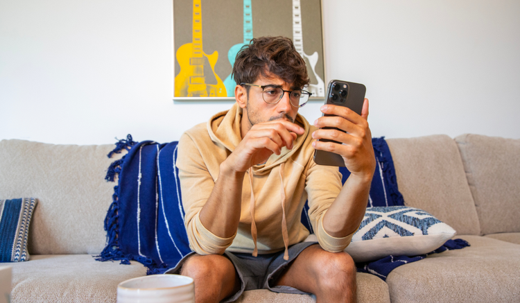 An image of a worried man in his living room, sitting on the couch. He is looking at his phone.