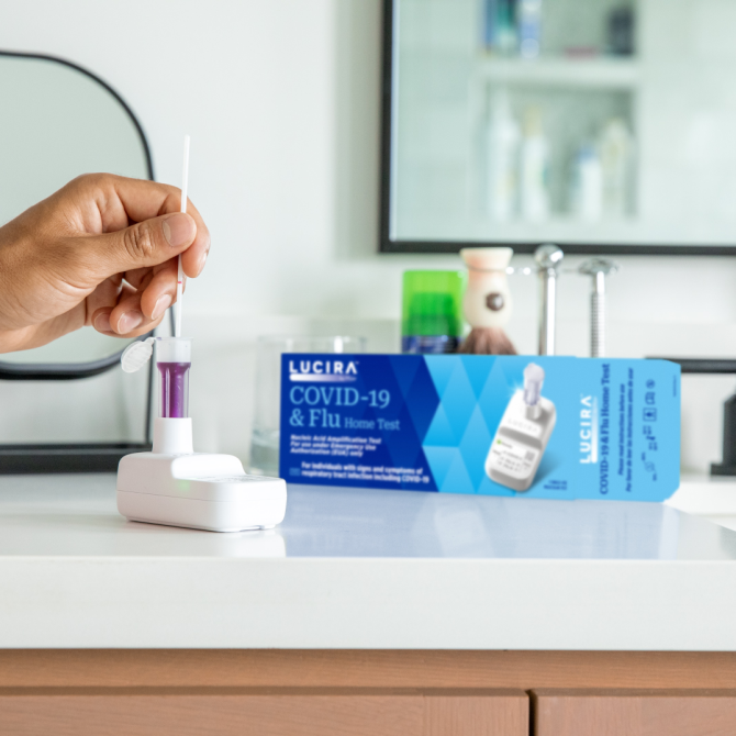 A hand comes in and uses the LUCIRA® by Pfizer COVID-19 & Flu Home Test with the packaging sitting on the right on top of the counter.