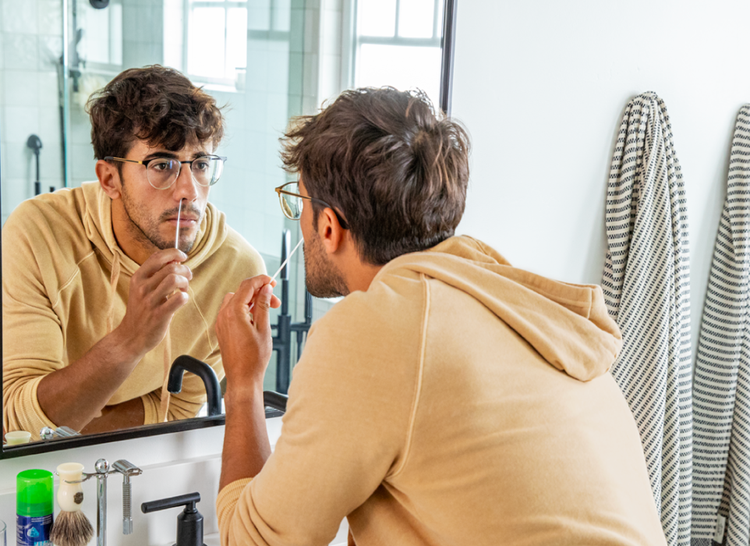 An image of a man with glasses as he administers a nasal swab in his bathroom while looking in the mirror.