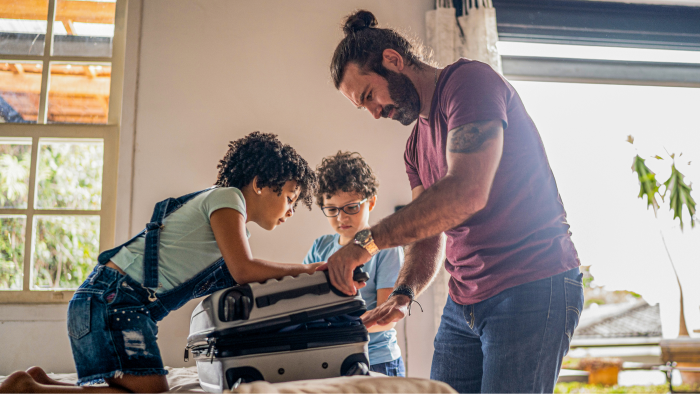 An image of a father helping his two sons pack a suitcase in a light and airy room of a house.