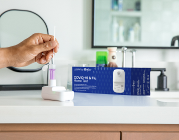 A hand comes in and uses the LUCIRA® by Pfizer COVID-19 & Flu Home Test with the packaging sitting on the right on top of the counter.