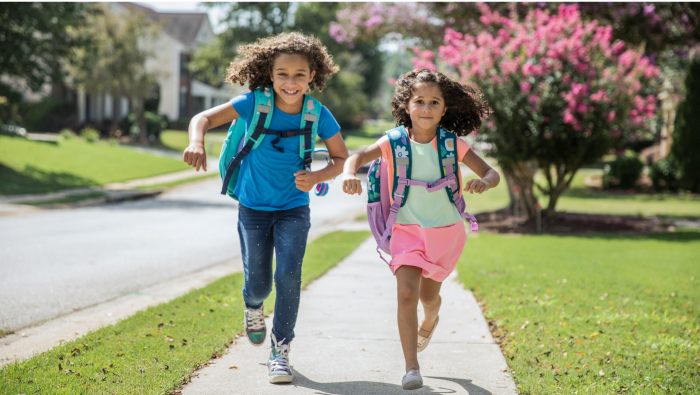 An image of two happy school-aged girls with backpacks running on a sidewalk of an idyllic neighborhood.