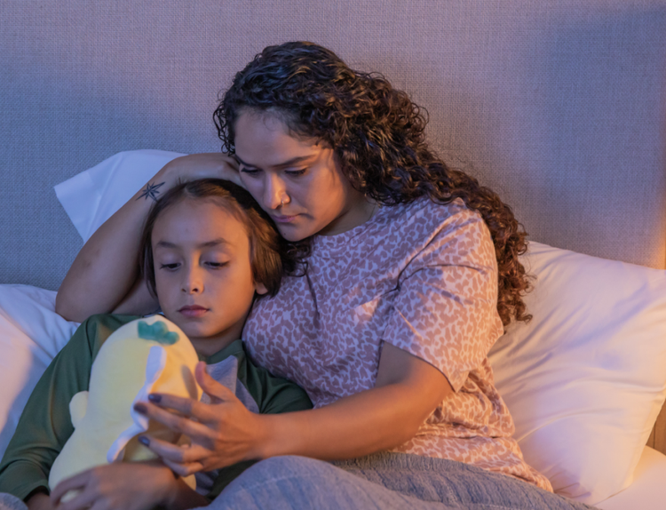 An image of a concerned mom lying in bed with her child, as they look at a stuffed animal.