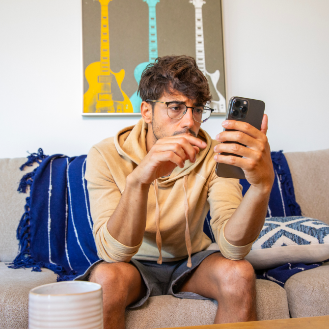 An image of a worried man in his living room, sitting on the couch. He is looking at his phone.