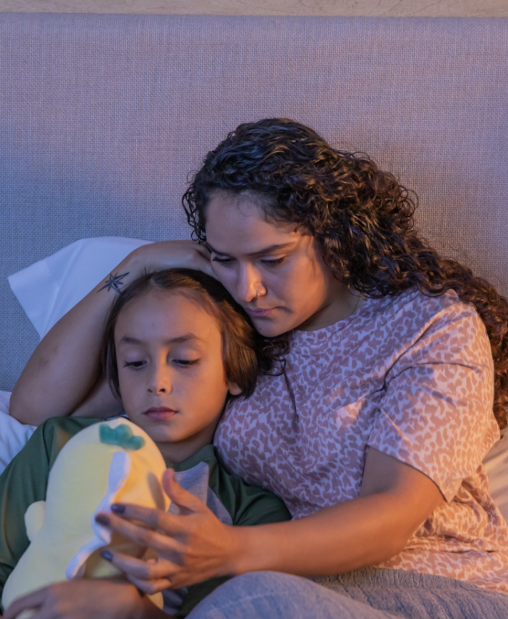 An image of a concerned mom lying in bed with her child, as they look at a stuffed animal.