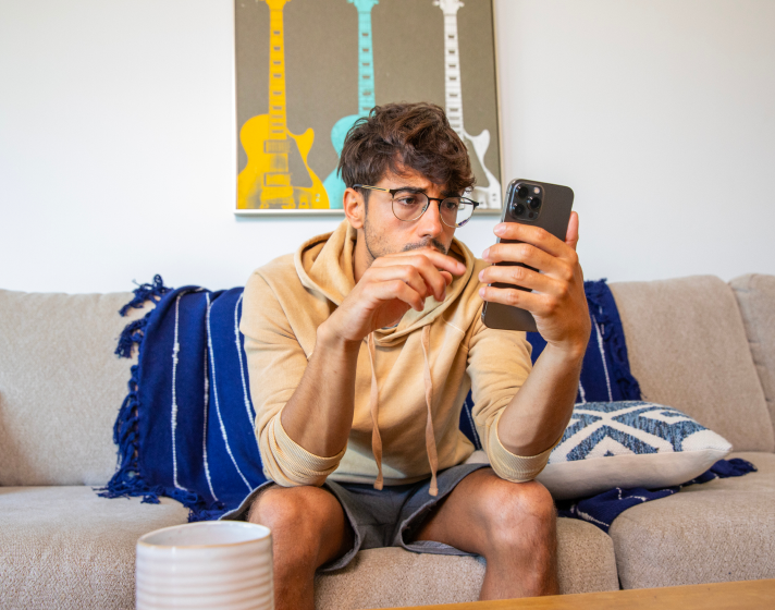 An image of a worried man in his living room, sitting on the couch. He is looking at his phone.