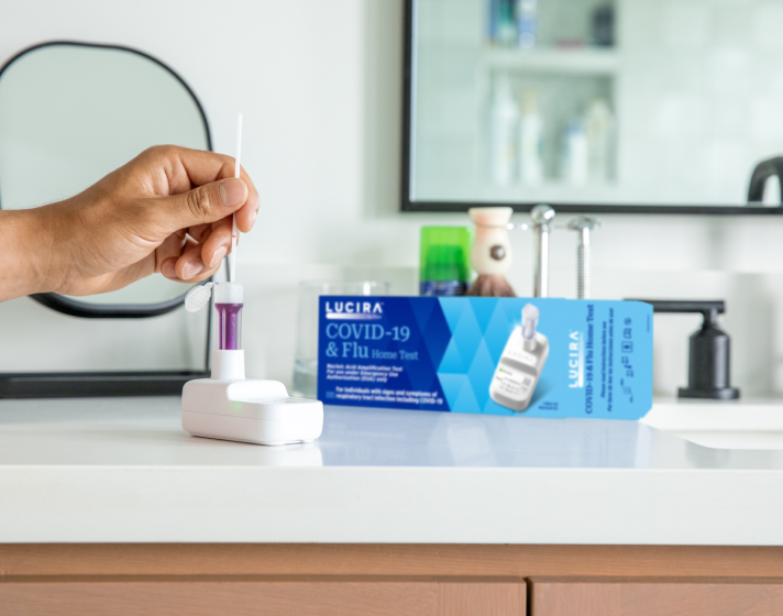 A hand comes in and uses the LUCIRA® by Pfizer COVID-19 & Flu Home Test with the packaging sitting on the right on top of the counter.