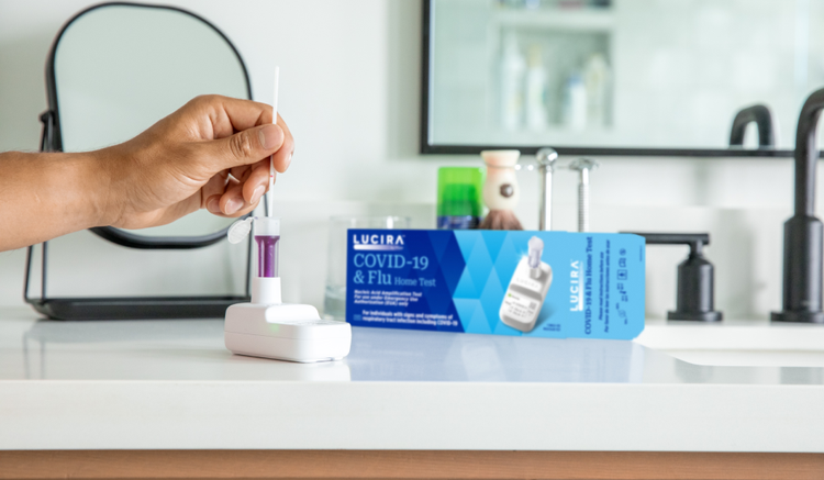 A hand comes in and uses the LUCIRA® by Pfizer COVID-19 & Flu Home Test with the packaging sitting on the right on top of the counter.