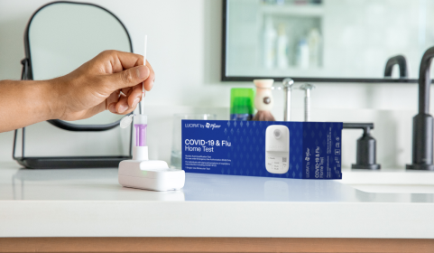 A hand comes in and uses the LUCIRA® by Pfizer COVID-19 & Flu Home Test with the packaging sitting on the right on top of the counter.