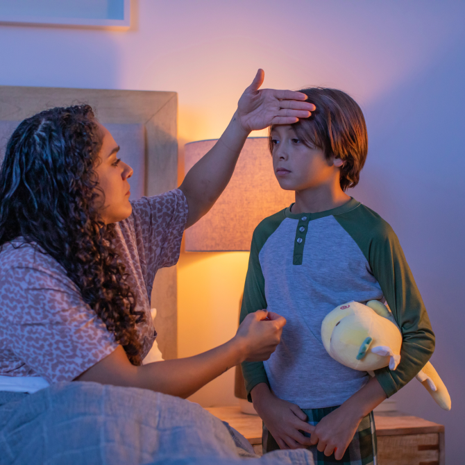 An image of a mom sitting on the bed as her child stands in front of her. Her hand is on the child's head to check his temperature.