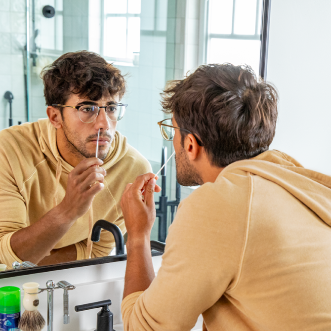 An image of a man with glasses as he administers a nasal swab in his bathroom while looking in the mirror.