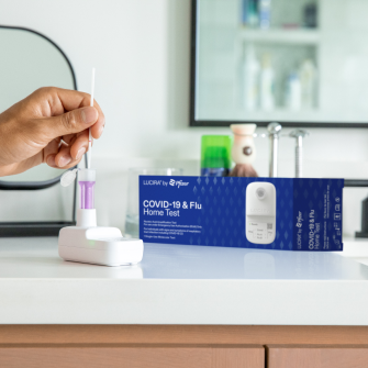A hand comes in and uses the LUCIRA® by Pfizer COVID-19 & Flu Home Test with the packaging sitting on the right on top of the counter.